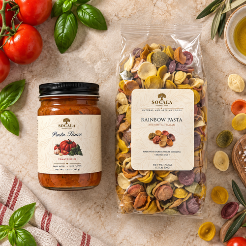 A jar of Socala tomato sauce and a large bag of Socala Rainbow Pasta are arranged on a textured stone surface with fresh tomatoes, basil leaves, and olive oil, creating an appealing food product presentation.