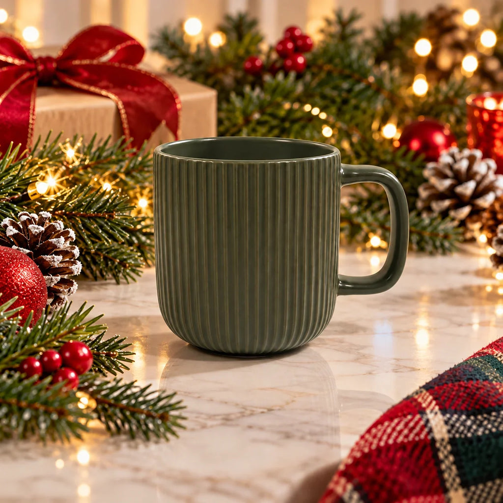 A ribbed ceramic mug in a muted sage green color, set on a marble surface surrounded by festive Christmas decorations including pine branches, red ornaments, a gift box with a red bow, and warm string lights.