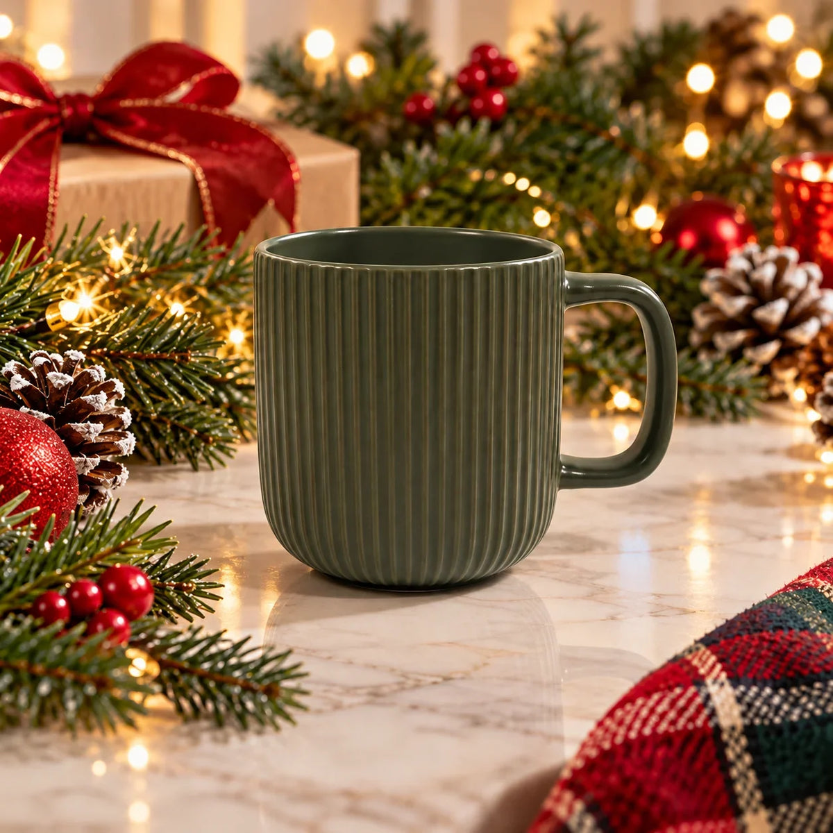 A ribbed ceramic mug in a muted sage green color, set on a marble surface surrounded by festive Christmas decorations including pine branches, red ornaments, a gift box with a red bow, and warm string lights.