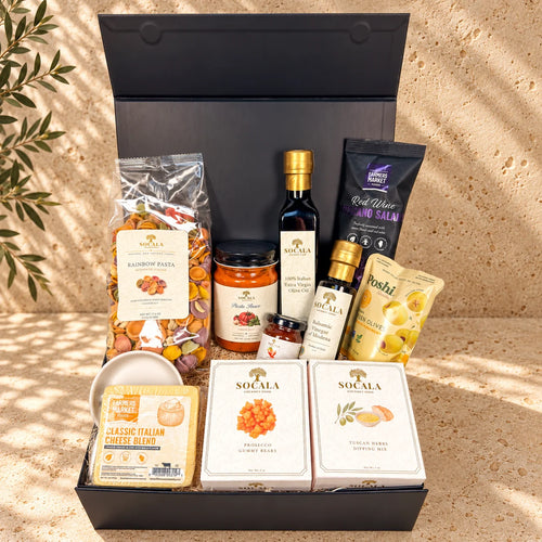 A gourmet gift hamper from Socala, featuring an assortment of Italian foods including pasta, salami, olive oil, cheese, and crackers, neatly arranged in an open black gift box on a sandy surface with leaf shadows.