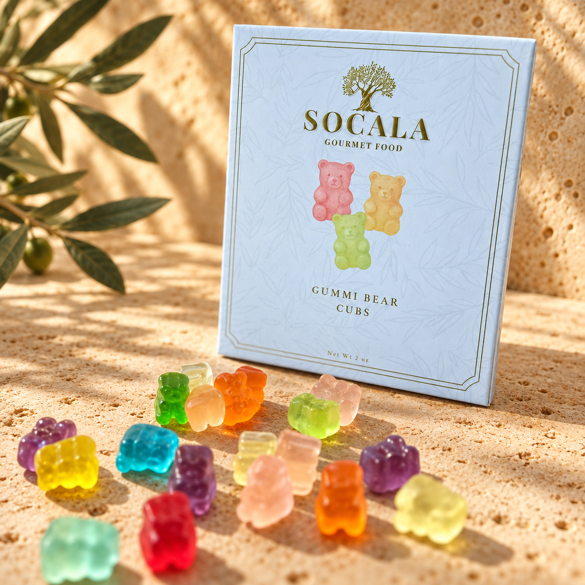A box of Socala Gourmet Food Gummi Bear Cubes stands on a sandy surface with several colorful gummy bears scattered in front. The box is pale blue with a tree logo and the words 'SOCALA GOURMET FOOD' and 'GUMMI BEAR CUBES.