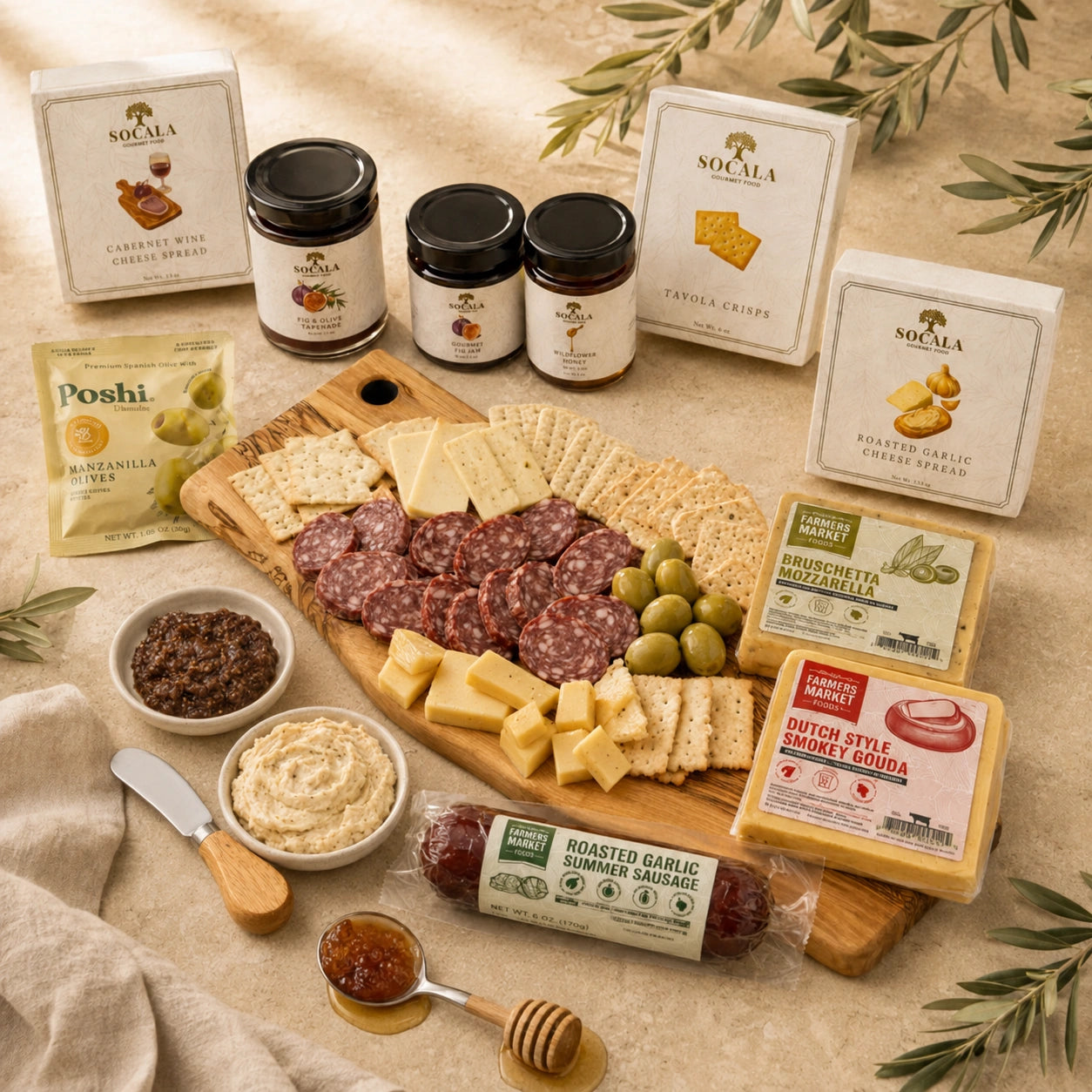 A styled charcuterie and cheese spread arranged on a wooden board with crackers, sliced salami, olives, cheese cubes, dips, jars, and branded boxes on a beige surface with olive branches around it.