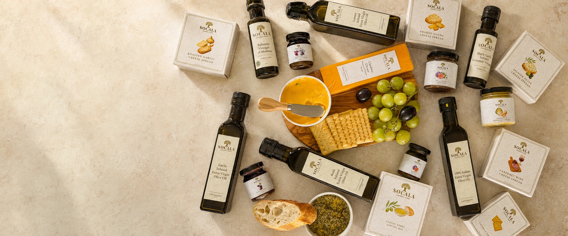An overhead flat-lay of a gourmet food and beverage assortment from the brand Socala, featuring olive oil bottles, honey pots, crackers, grapes, and cheese on a light beige stone surface.
