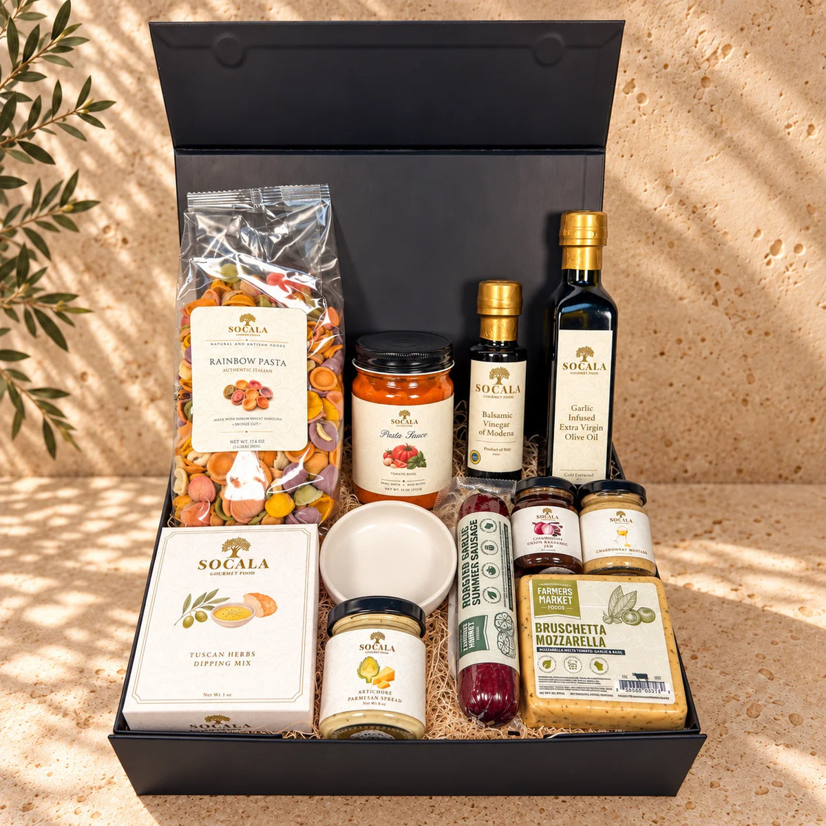 A curated gourmet gift hamper in an open black box, featuring food products from Micalà and Socala. The assortment includes a bag of rainbow pasta, a jar of fruit jam, a bottle of garlic olive oil, a box of sunflower seeds, jars of spreads, and a small bowl, arranged on a textured beige stone surface with dappled sunlight and leaf shadows.