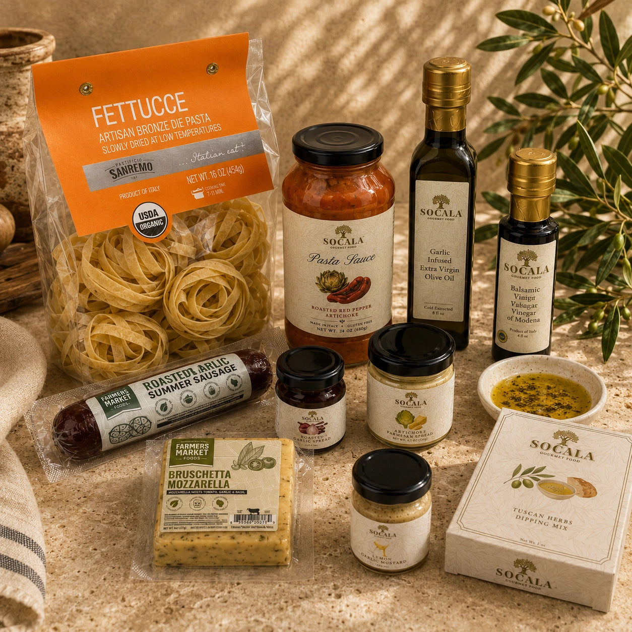 A styled flat-lay of a Socala family dining set on a beige stone surface, featuring fettuccine pasta, roasted summer sausage, bruschetta mozzarella, olive oil, vinegar, honey, and a honey pot holder box.