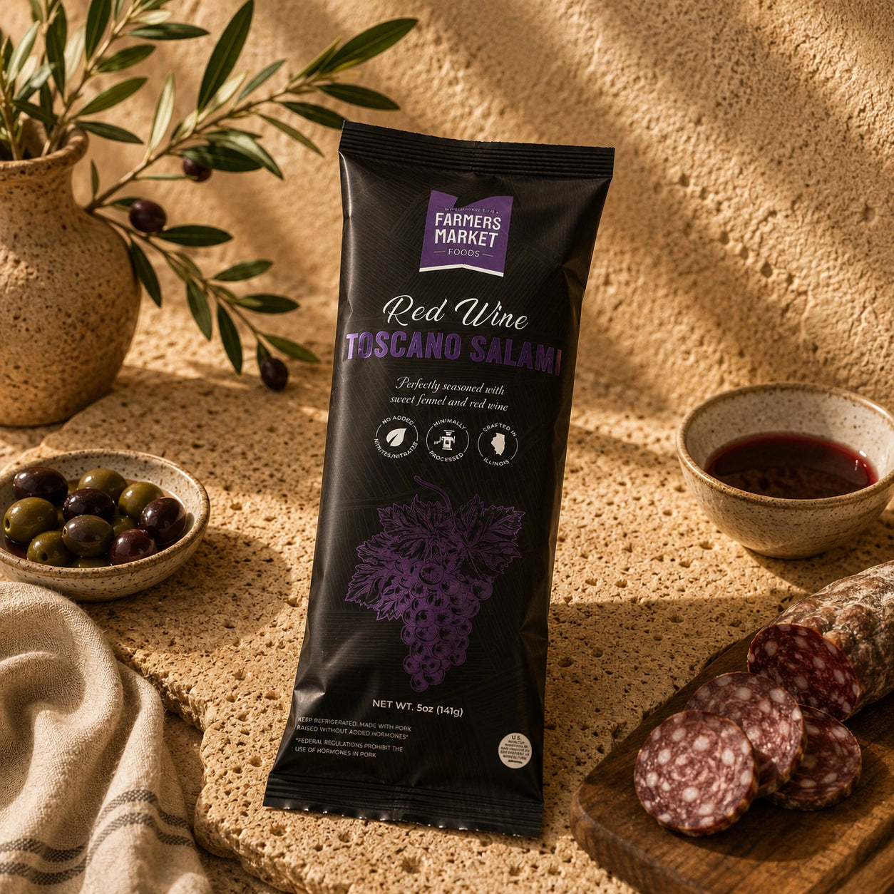 A package of Farmers Market Red Wine Toscano Salami stands upright on a textured stone surface, surrounded by olive branches, olives, a bowl of red wine, and sliced salami on a wooden board.