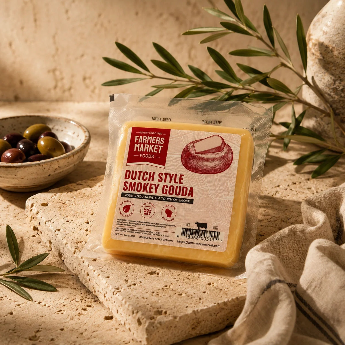 A packaged wedge of cheese labeled 'Dutch Style Smokey Gouda' by Farmers Market Foods, placed on a stone slab with olive branches, olives, and a linen cloth in a warm rustic setting.