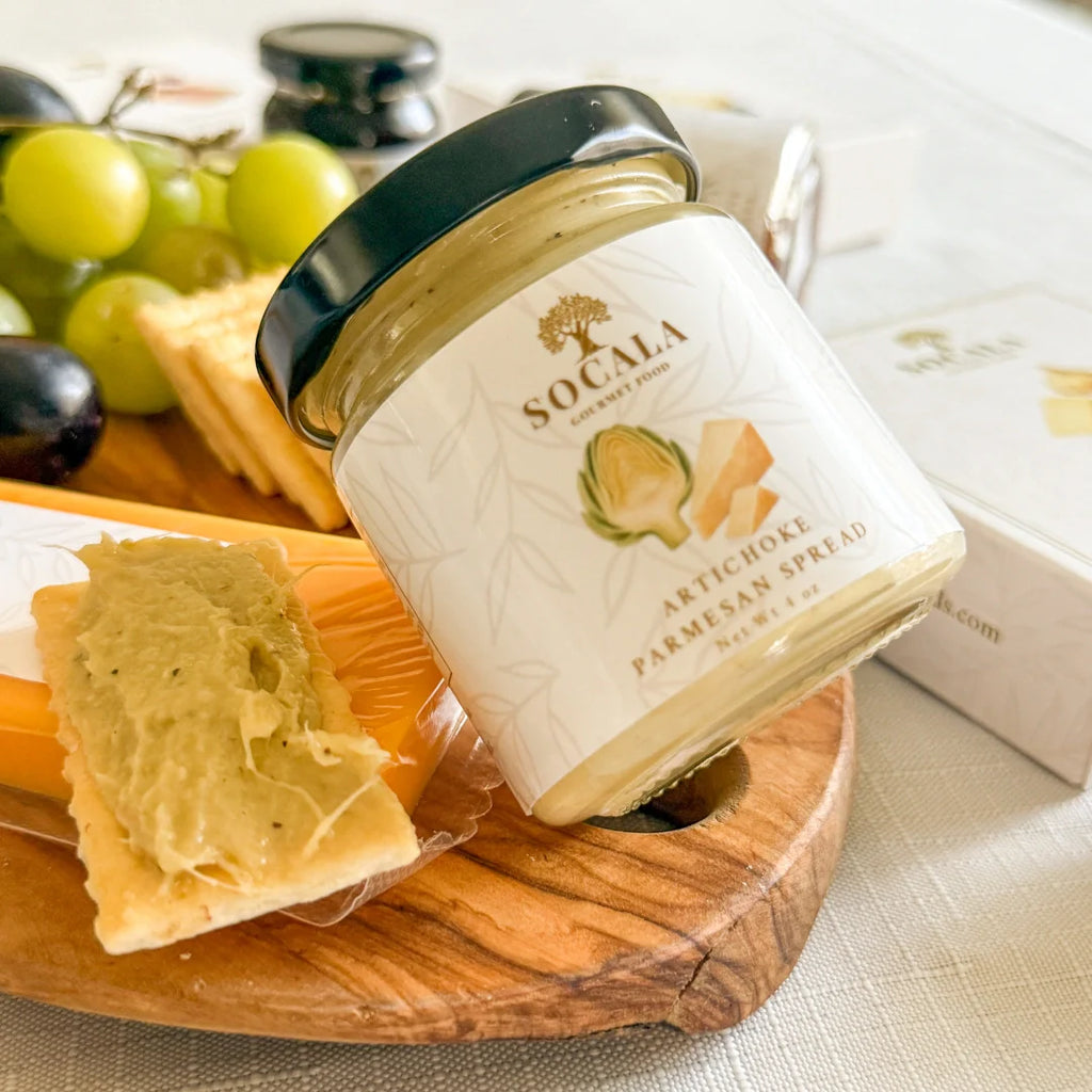 Jar of Socala artichoke Parmesan spread on a wooden board with crackers and grapes.