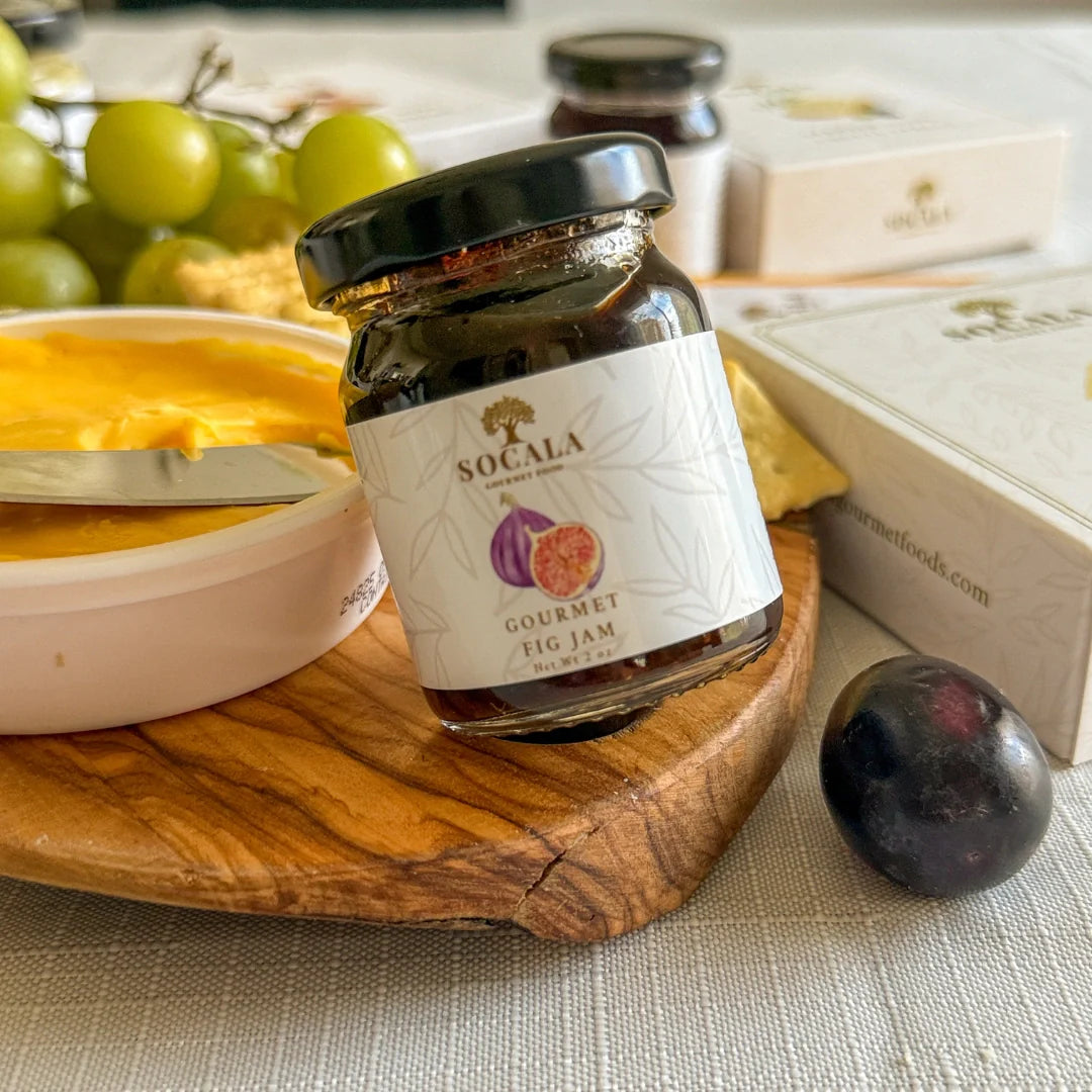 Jar of Socala Gourmet Fig Jam on a wooden board with fruits and cheese in the background.