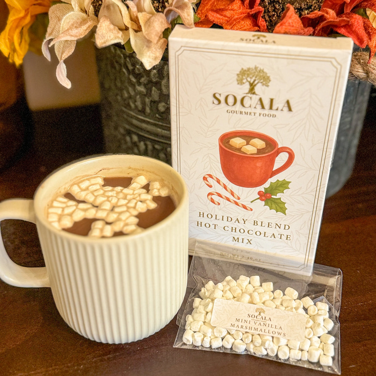 Socala holiday blend hot chocolate mix with a mug of hot chocolate and marshmallows on a wooden surface.