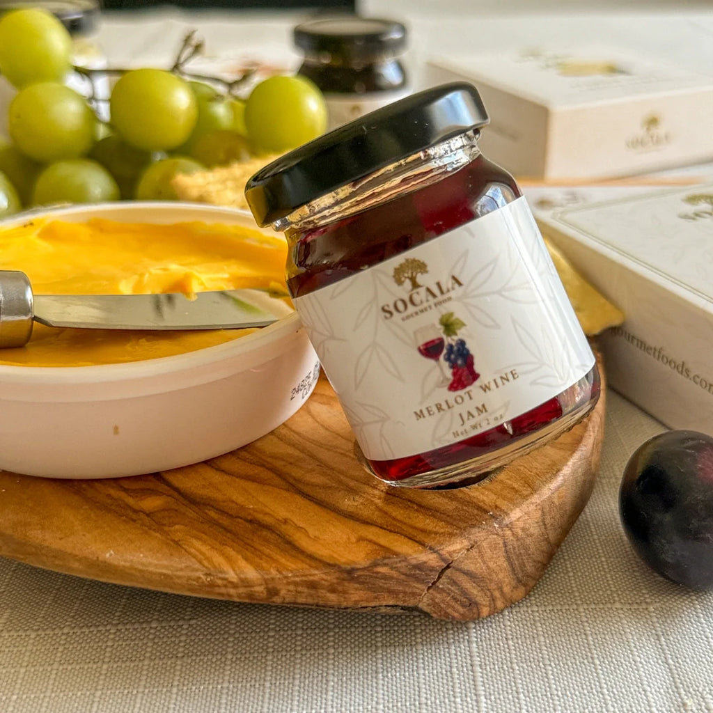 Jar of Socala Merlot Wine Jam on a wooden board with cheese and grapes in the background.