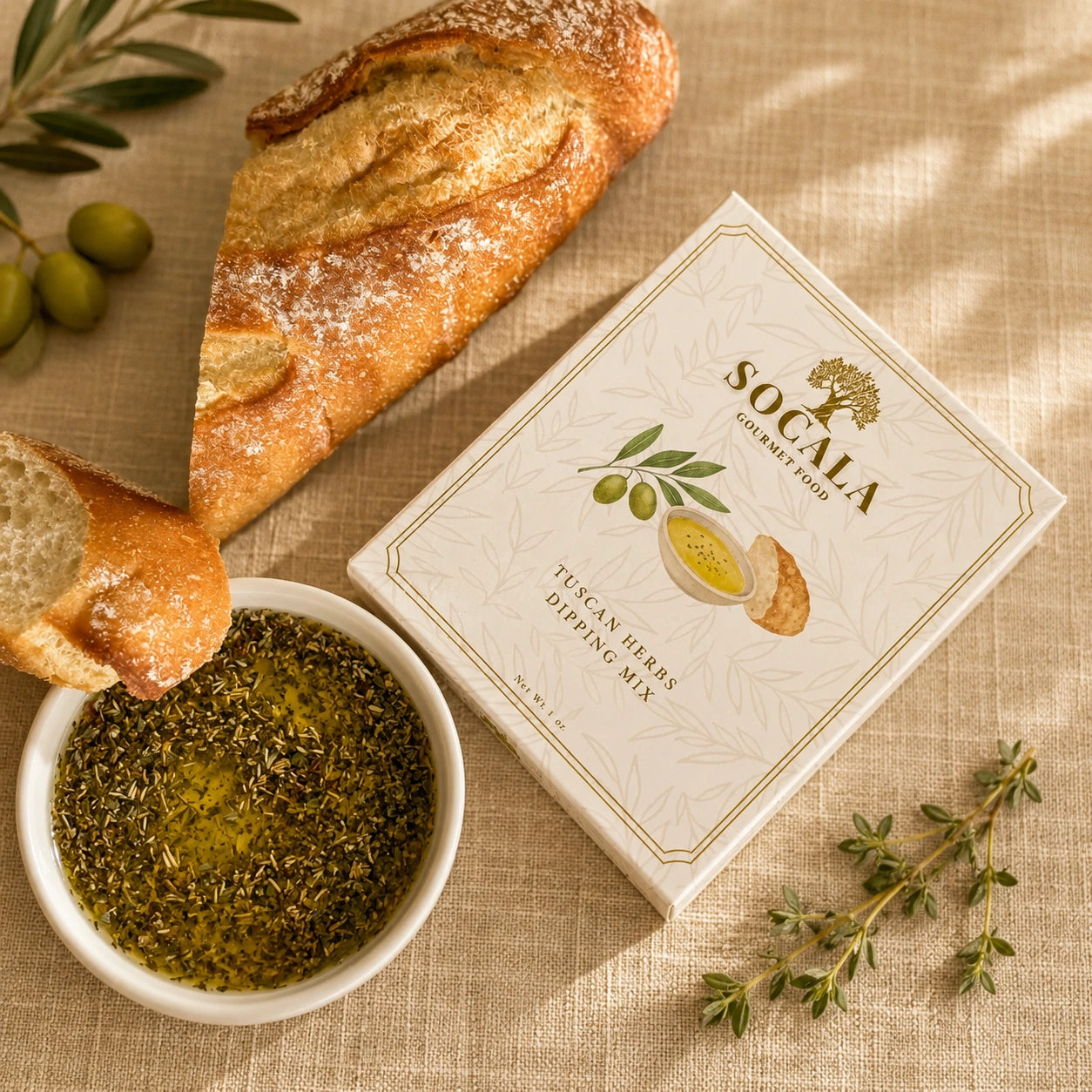 A boxed package of Socala Olive Oil & Herb Dipping Mix placed on a linen surface beside a bowl of herb-dusted dip, sliced rustic bread, and olives. The box is cream-colored with olive-green and yellow illustrations and the brand name Socala clearly visible.